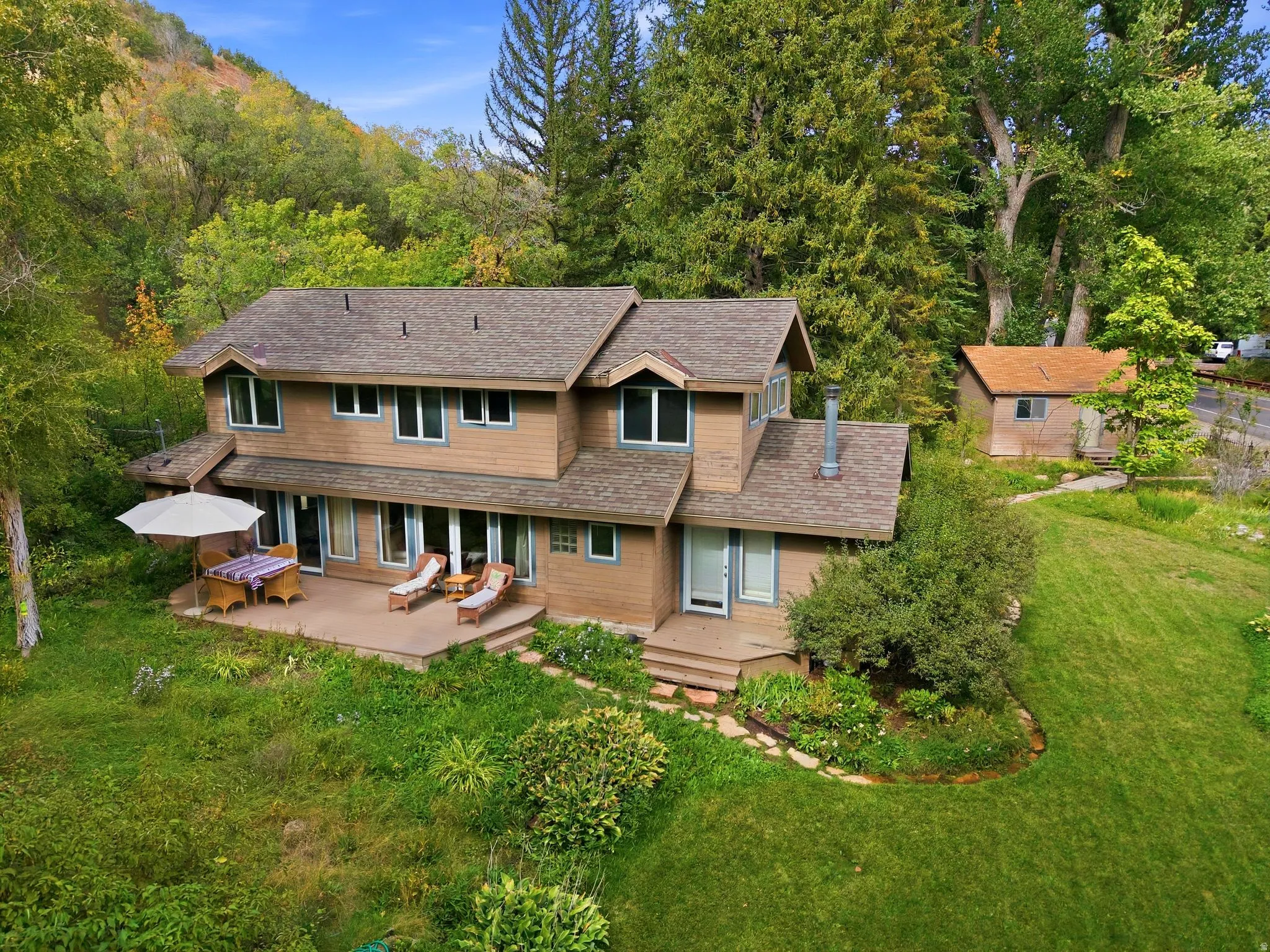 Multi-level residence with a wood-sided exterior, a spacious rear patio, and a separate outbuilding, all set on a landscaped lot with mature trees and a grassy expanse