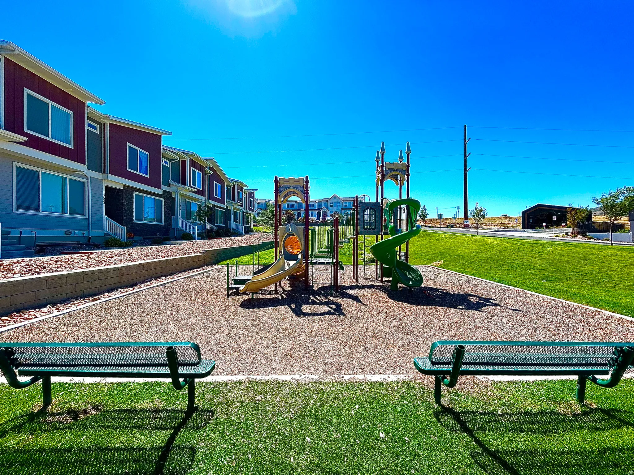 Community playground with multiple slides and climbing features, situated on a wood chip surface, with two park benches and a manicured lawn area