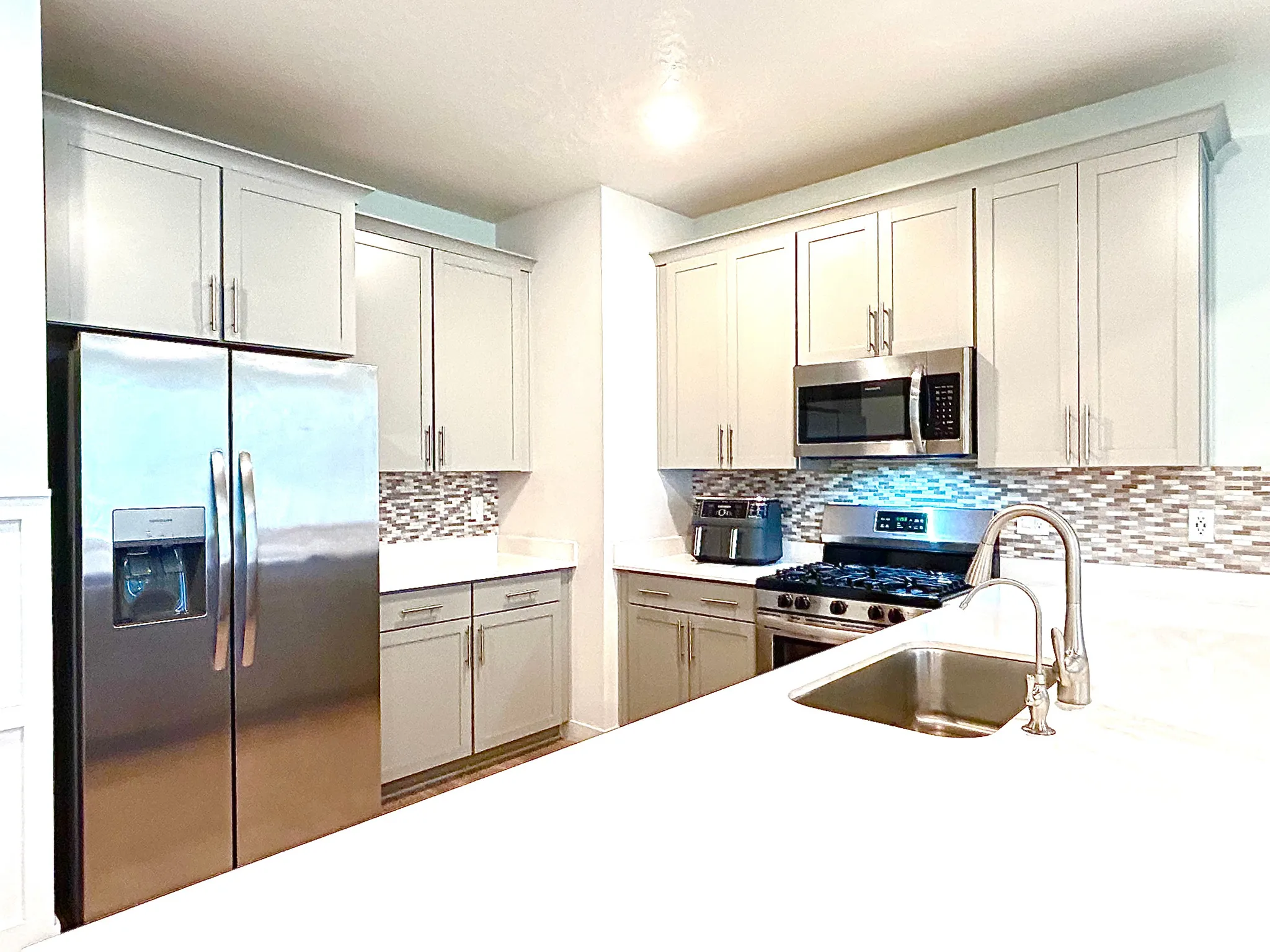 Stainless steel appliances, light-toned cabinetry, solid surface countertops, mosaic tile backsplash, and a stainless steel sink with a gooseneck faucet