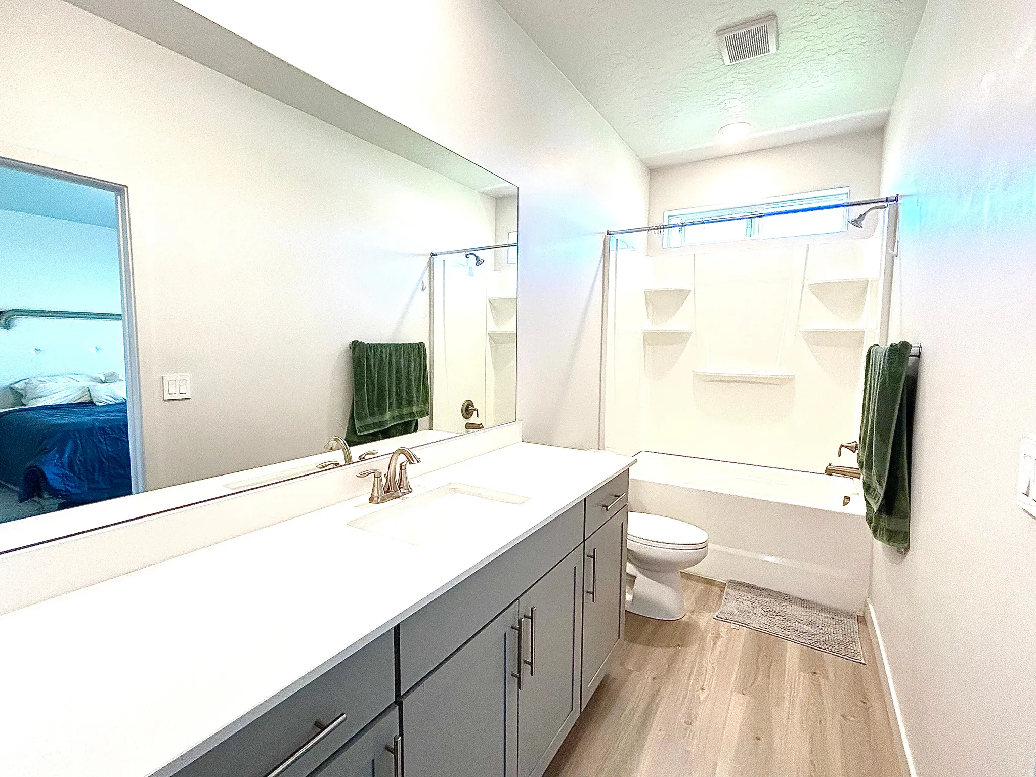 Dual vanity with integrated sinks, light-toned wood-finish flooring, a full-sized bathtub with an overhead shower, a window above the bathtub, and built-in vanity cabinetry