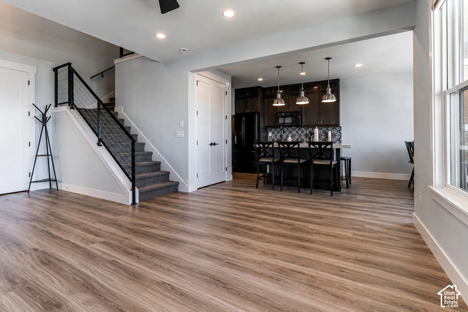 Open-concept living space featuring wood-finish flooring, a kitchen island with pendant lighting, dark wood cabinetry, and a carpeted staircase with cable railing