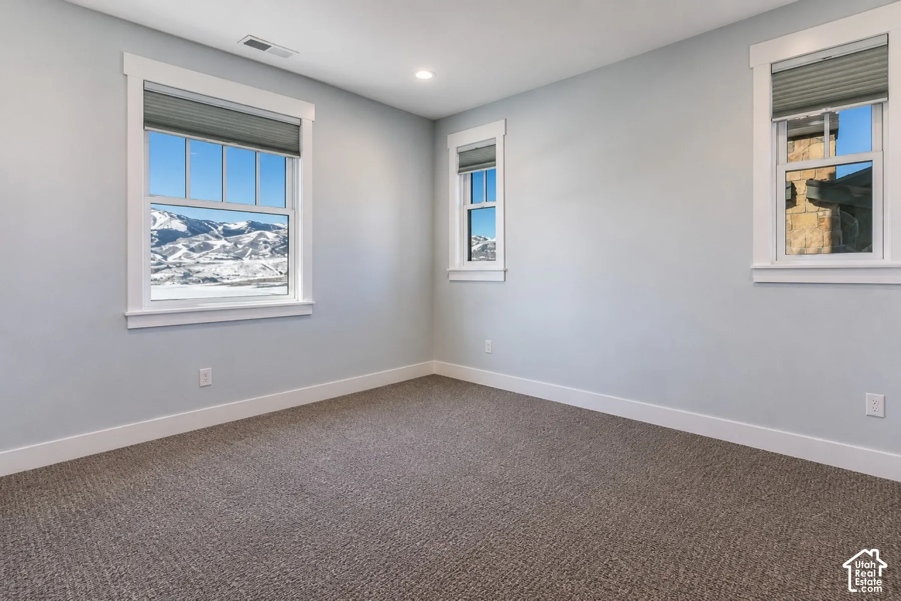 Light gray wall paint, recessed ceiling lighting, three windows with white trim, gray textured carpet, and white baseboards