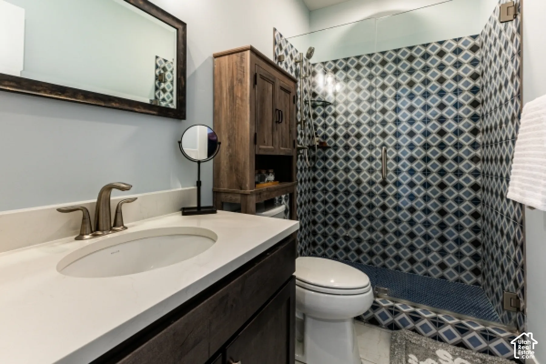 Vanity with an integrated sink and light-toned countertop, framed mirror, brushed nickel faucet, and glass-enclosed shower with patterned tile surround