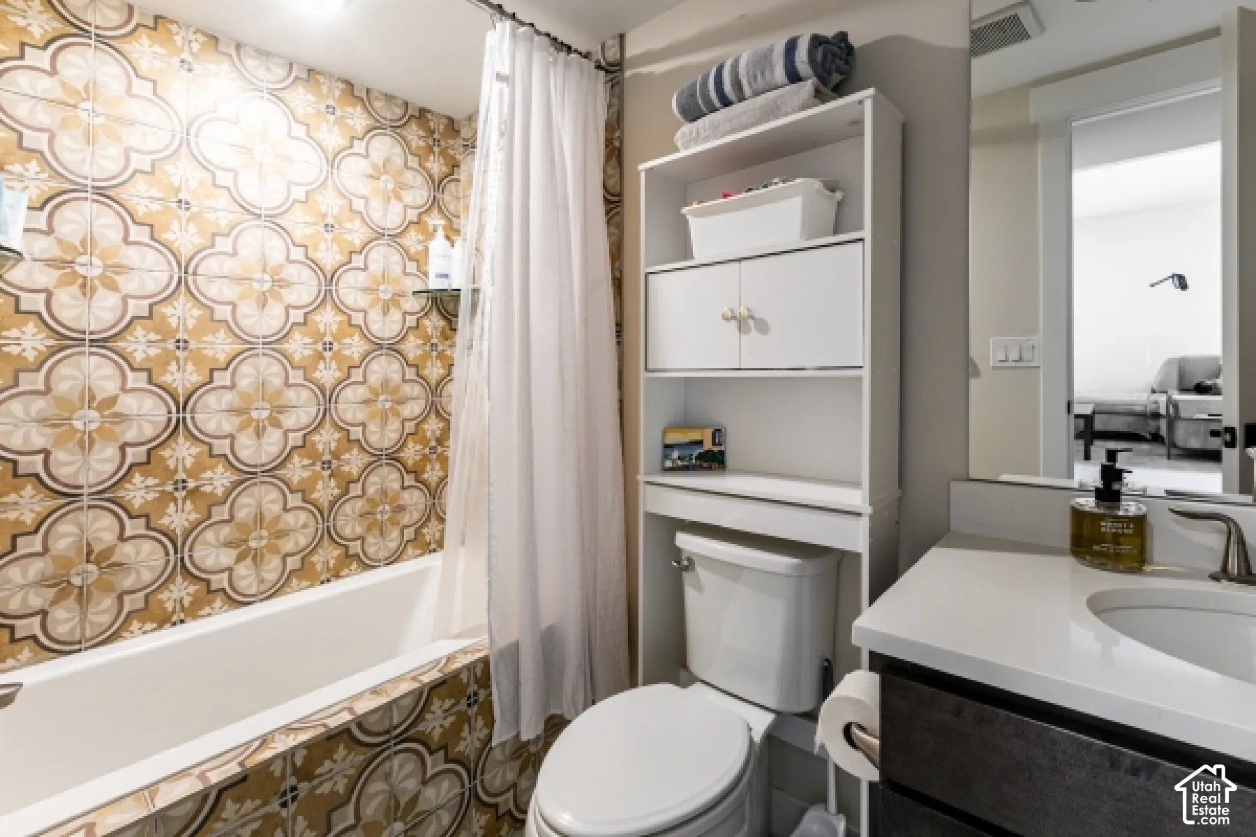 Bathtub with decorative patterned tile surround, white vanity with integrated sink, and white storage shelving over the toilet