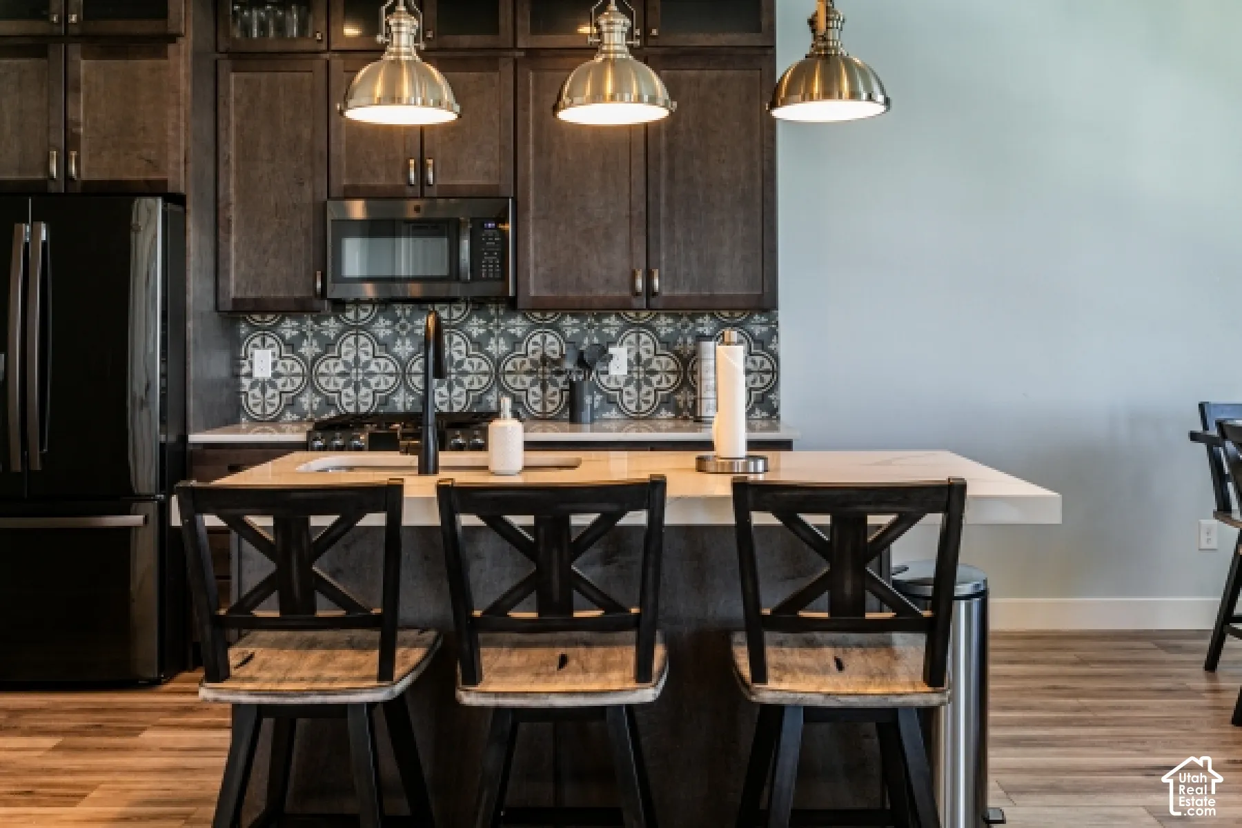 Wood-finish flooring, dark wood cabinetry with glass accents, decorative tile backsplash, white countertop, and stainless steel appliances