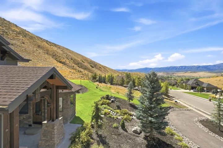 Stone facade with wood beam accents, shingle roofing, sloped landscaping with manicured lawn, paved driveway