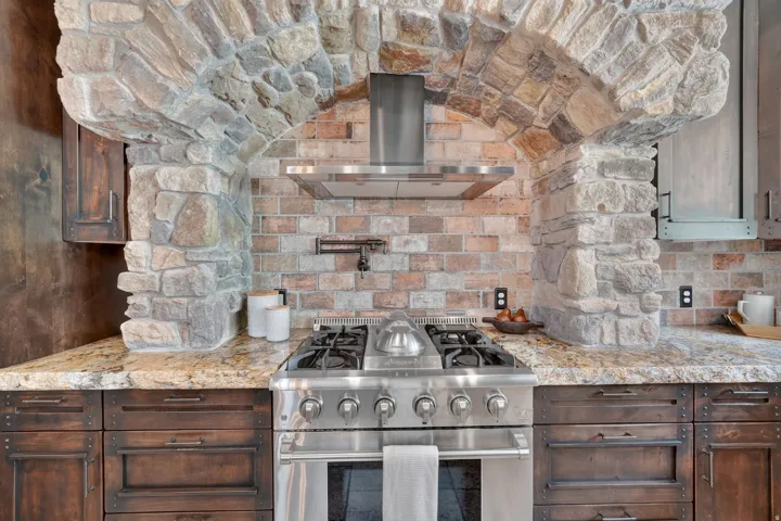 Stone archway with a brick backsplash, stainless steel range, built-in range hood, and wood cabinetry