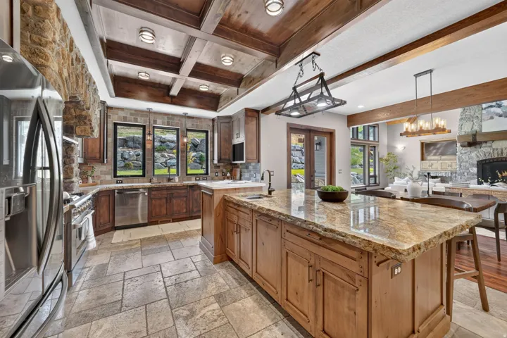 Gourmet kitchen with natural stone flooring, custom wood cabinetry, stainless steel appliances, a large center island with a natural stone countertop, and a wooden coffered ceiling