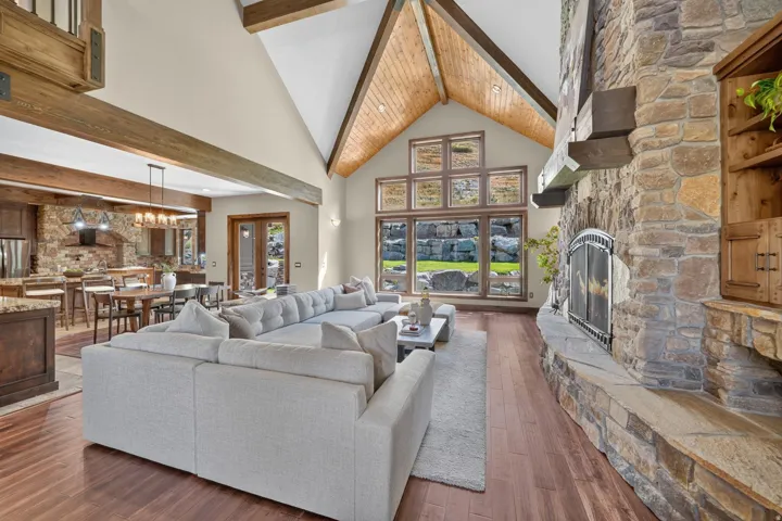 Vaulted ceiling with exposed wood beams, stone fireplace, wood-finish flooring, large picture windows, and double French doors