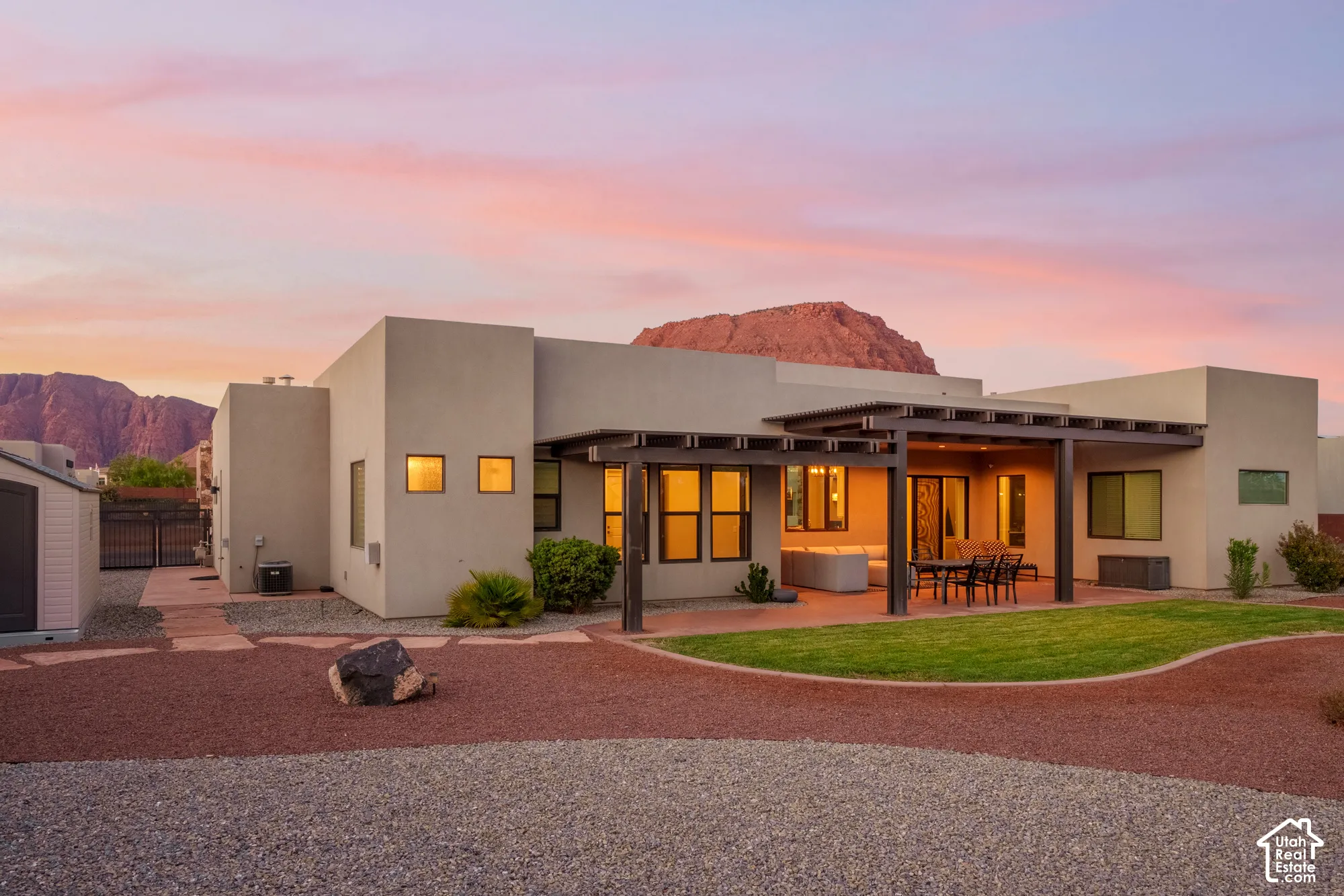 Stucco exterior with a covered patio featuring a slatted pergola, a landscaped yard with green lawn and gravel, and a detached storage shed