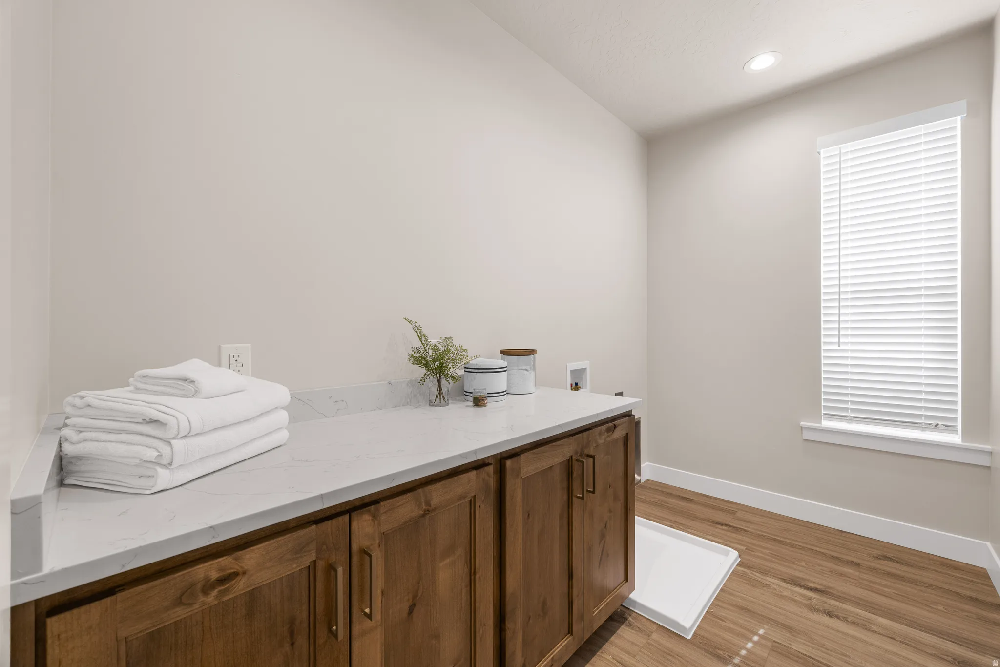Wood-finish flooring, white walls, wood cabinetry with modern hardware, and a window with blinds