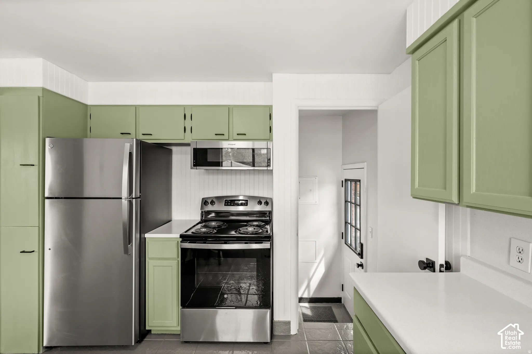 Stainless steel appliances, white countertops, green cabinetry, beadboard wall paneling, and tile flooring