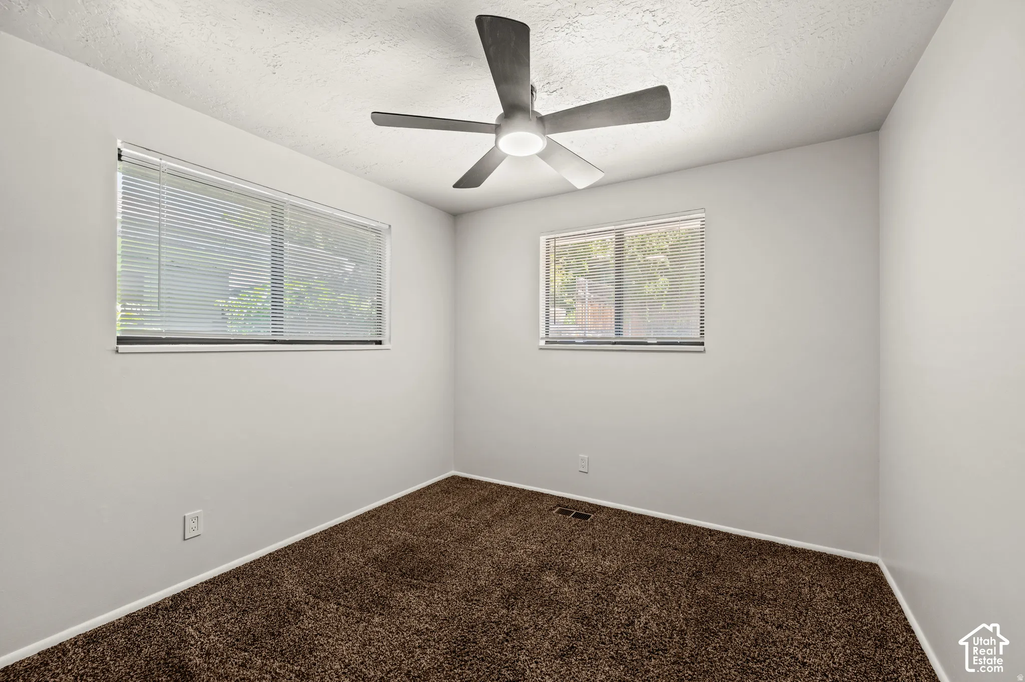 Neutral wall color, two windows, textured ceiling, ceiling fan with integrated lighting, and brown carpet flooring