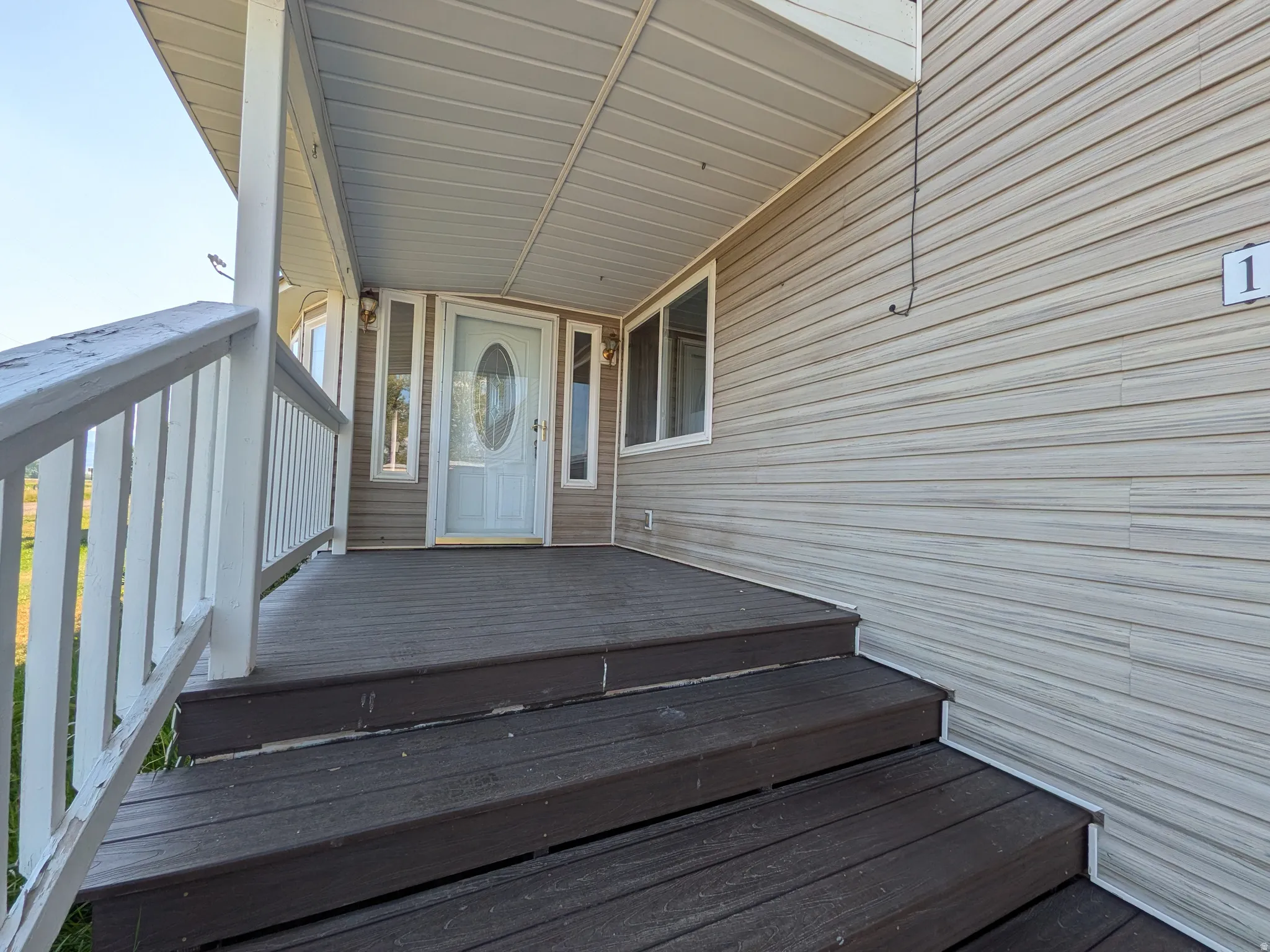 Covered front porch with wood-finish decking and white railing, featuring a white entry door with oval glass insert and flanking windows