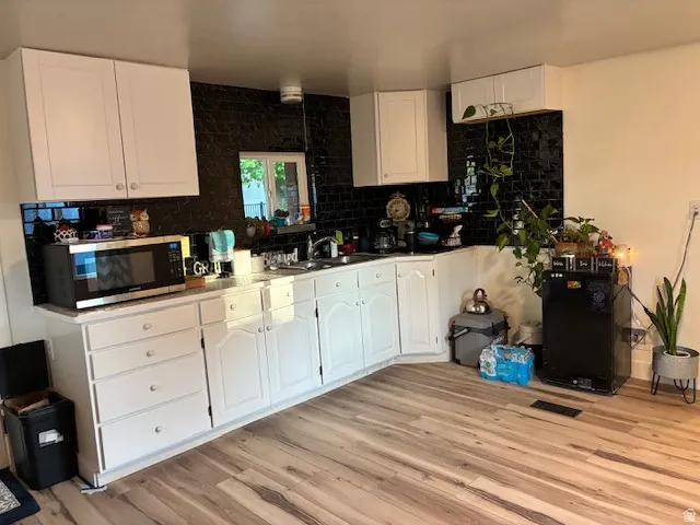 White cabinetry, black tile backsplash, wood-finish flooring, corner sink, and window