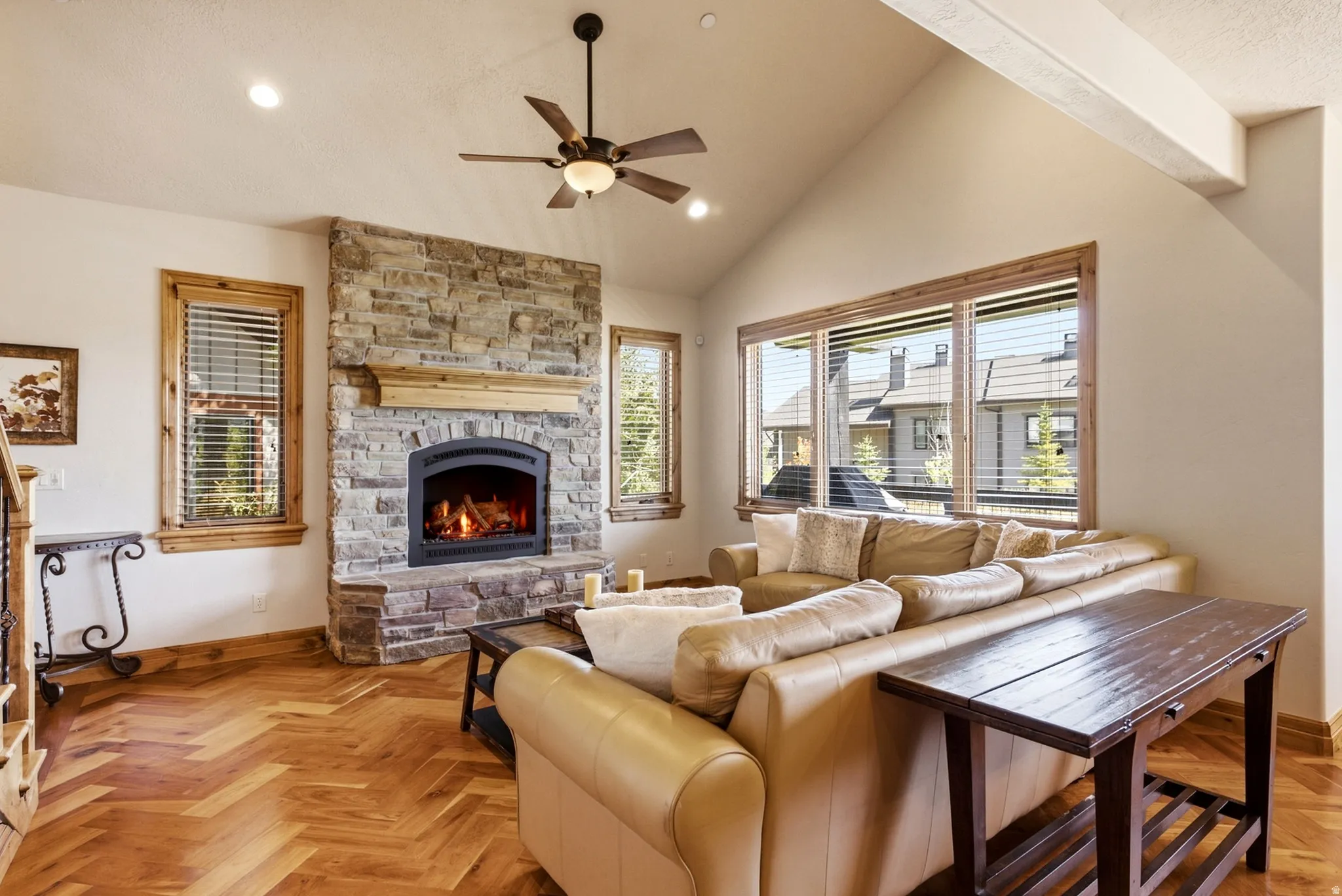 Vaulted ceiling with an installed ceiling fan, stone-clad fireplace, wood-finish flooring, and natural wood window trim
