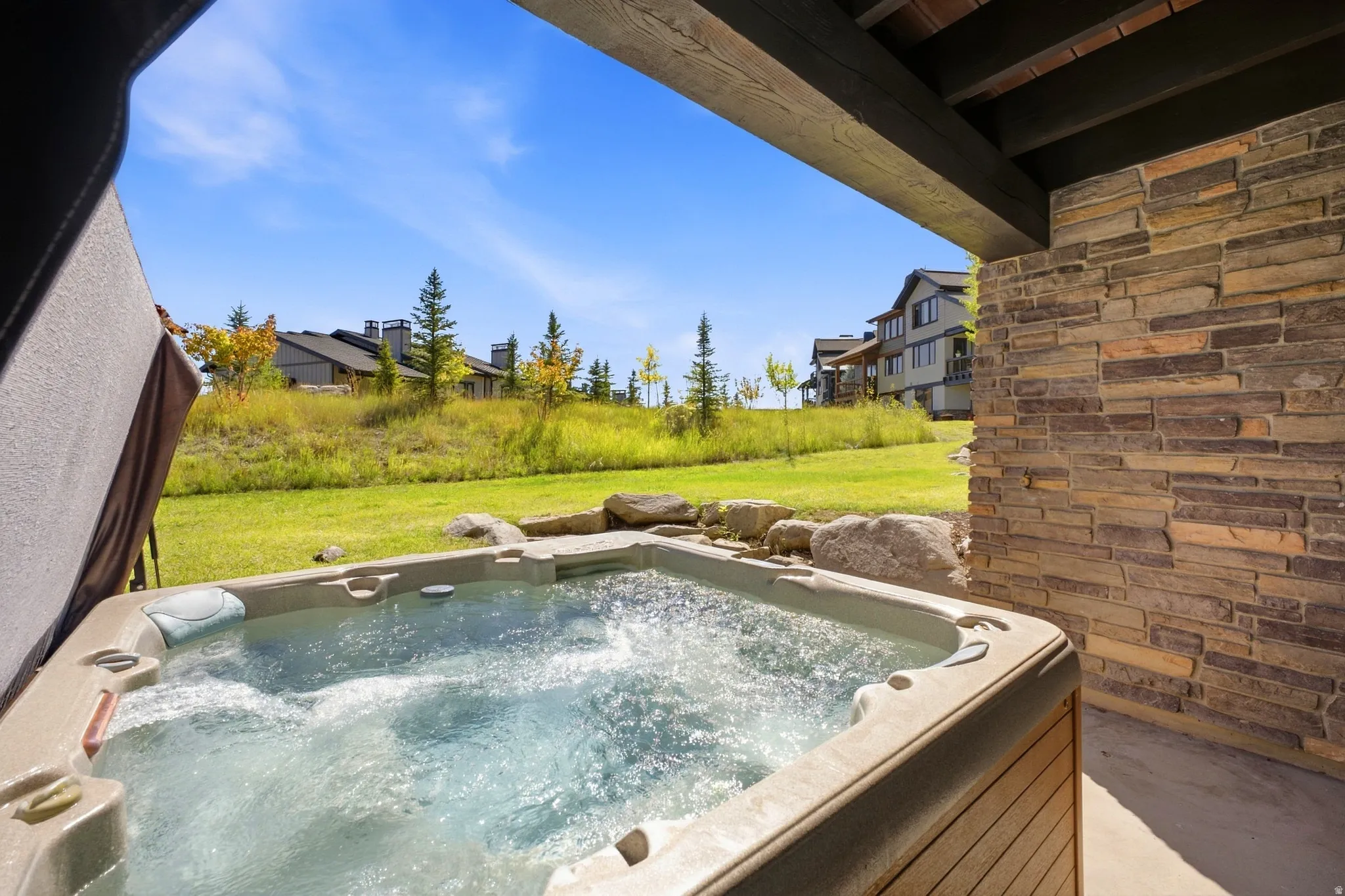 Built-in hot tub, stacked stone wall, concrete patio, exposed wood beam ceiling, and natural rock landscaping