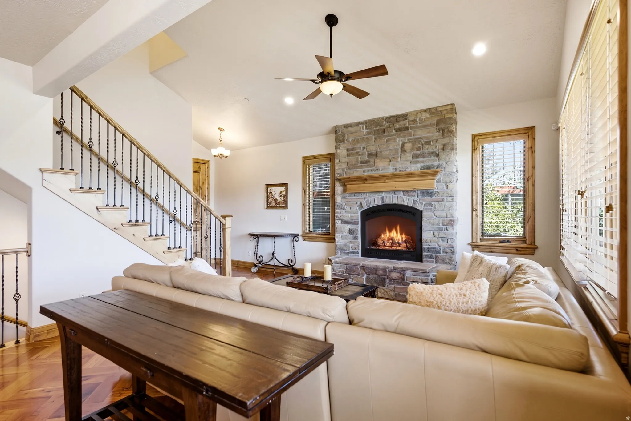 Stone-clad fireplace with wood mantel, ceiling fan with integrated lighting, wood-finish flooring, and a staircase with wrought iron balusters