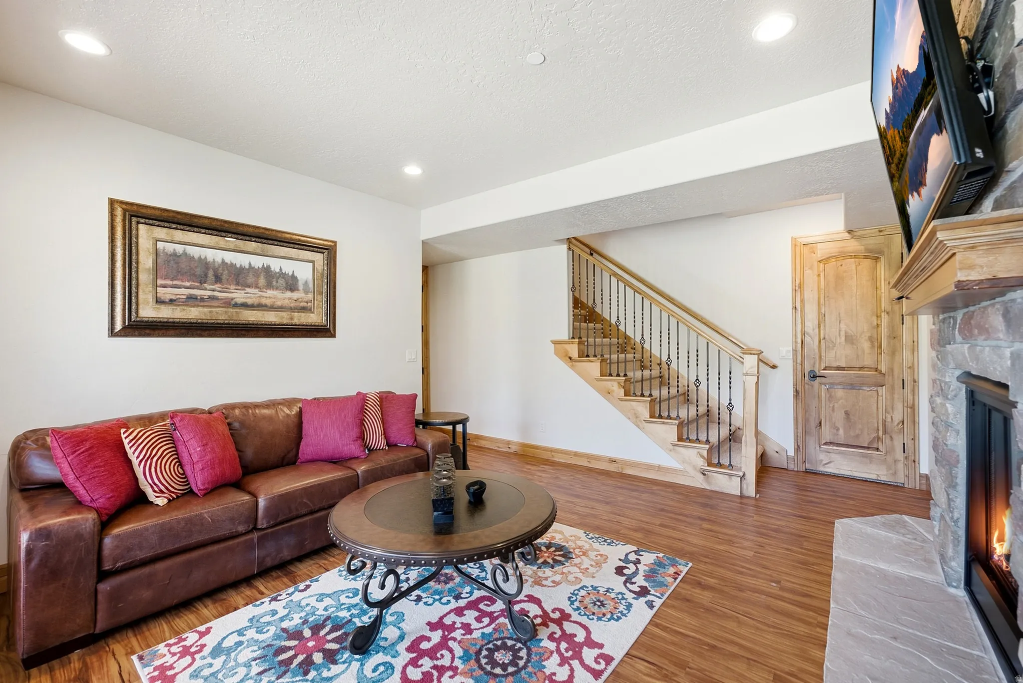 Stone-clad fireplace with wood mantel, wood-finish flooring, recessed lighting, and a staircase featuring wrought iron balusters