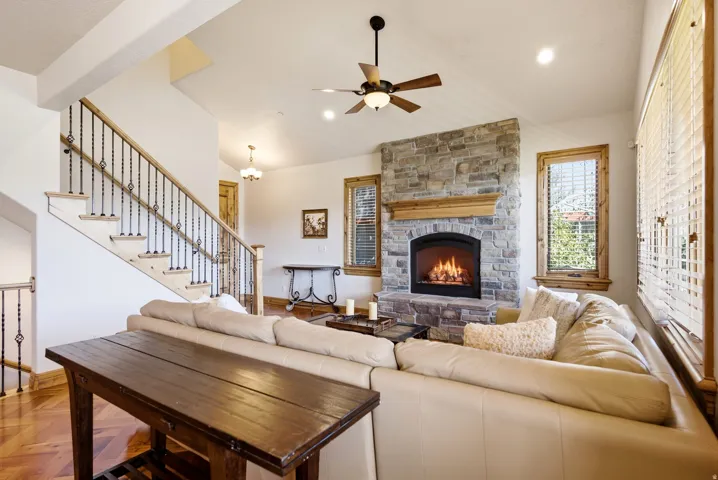 Stone-clad fireplace with wood mantel, ceiling fan with integrated lighting, wood-finish flooring, and a staircase with wrought iron balusters