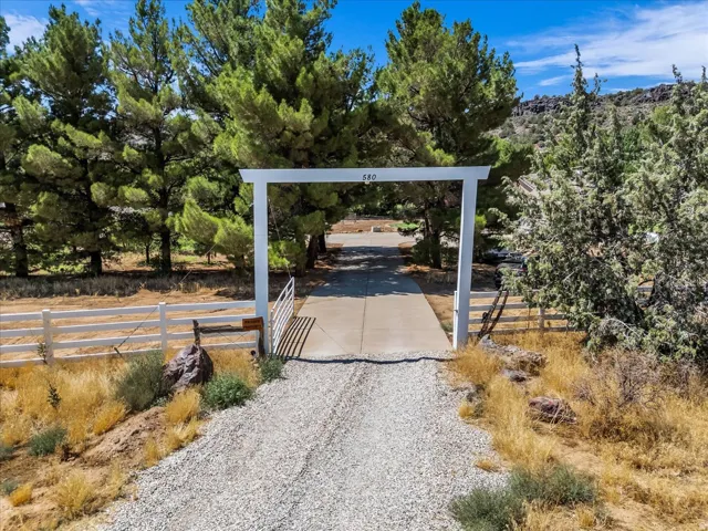 Gravel and concrete driveway with a white arched entry gate, bordered by a white fence and mature trees