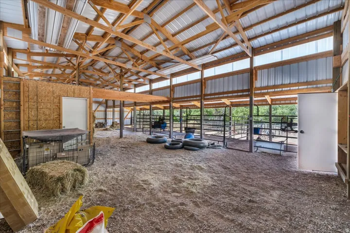 Spacious barn interior with exposed wood beam construction, corrugated metal roofing, multiple stalls with metal fencing, an interior room with a solid door, and natural light panels