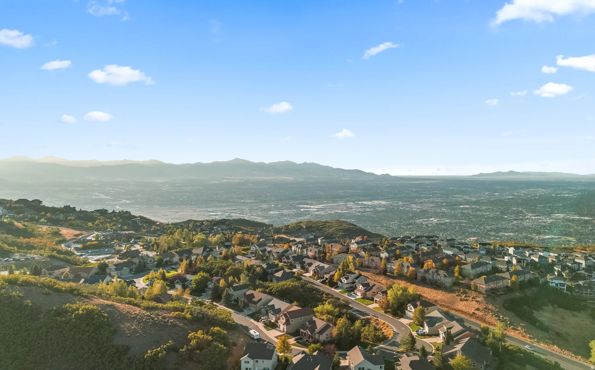 Expansive aerial perspective showcasing a residential community nestled into hillsides with distant mountain ranges and a sprawling metropolitan area visible in the background