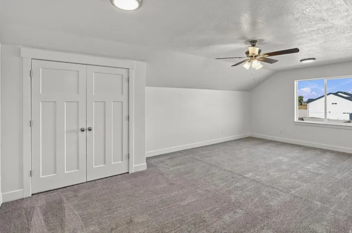 Spacious room featuring vaulted ceilings, ceiling fan, natural light from a large window, and light gray carpeting