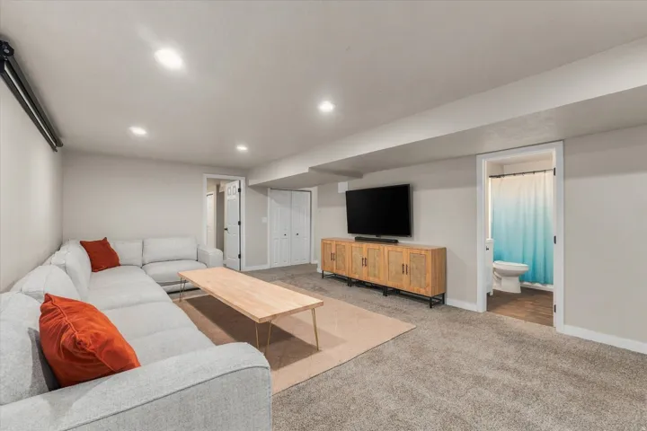 Recessed ceiling lighting, light gray wall paint, wood-finish media console, light gray carpeting, and an adjacent bathroom with a white toilet