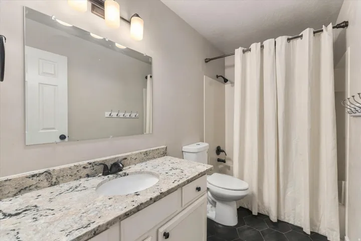 Vanity with a granite countertop and an undermount sink, dark hexagonal floor tiles, shower/tub combination with a dark-finish showerhead, and a three-light vanity fixture