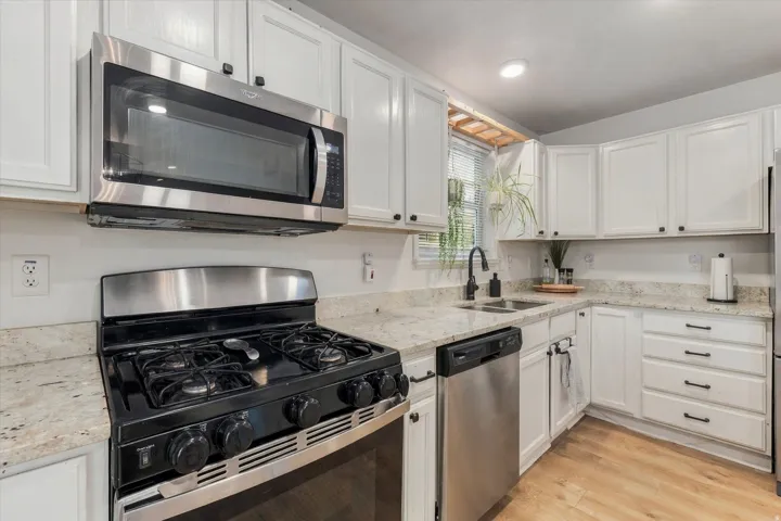 White shaker-style cabinetry with matte black hardware, stainless steel appliances, speckled stone countertops, undermount sink with a black gooseneck faucet, and wood-finish flooring