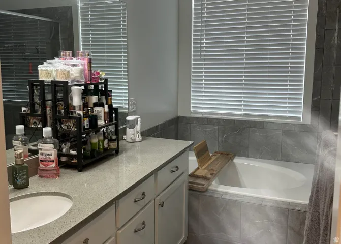 Built-in bathtub with tiled surround, adjacent window with blinds, and a vanity featuring a single sink and light-toned countertop