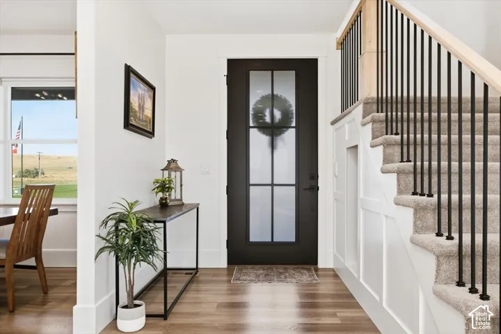 Wood-finish flooring, staircase with natural wood handrail and black metal balusters, board and batten wall detailing, and a black front door with frosted glass panels