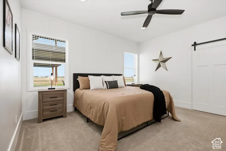 Plush carpet flooring, white paneled walls, a ceiling fan with integrated lighting, two windows with blinds, and a sliding barn door