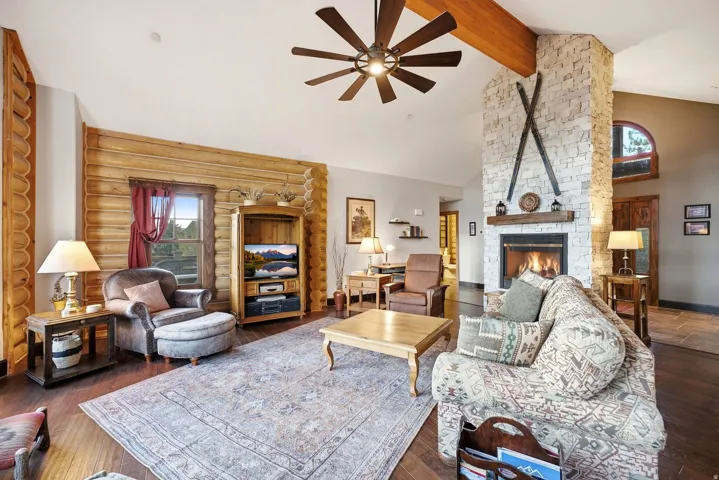 Stone-clad fireplace with wood mantel, wood-finish flooring, exposed wood beam, log wall accents, and a ceiling fan