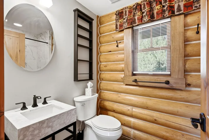 Rustic log cabin bathroom featuring horizontal wood-finish wall paneling, a marble-topped vanity with matte black hardware, an oval framed mirror, a window with wooden surround and horizontal blinds, white subway tile flooring, and vintage-style toilet