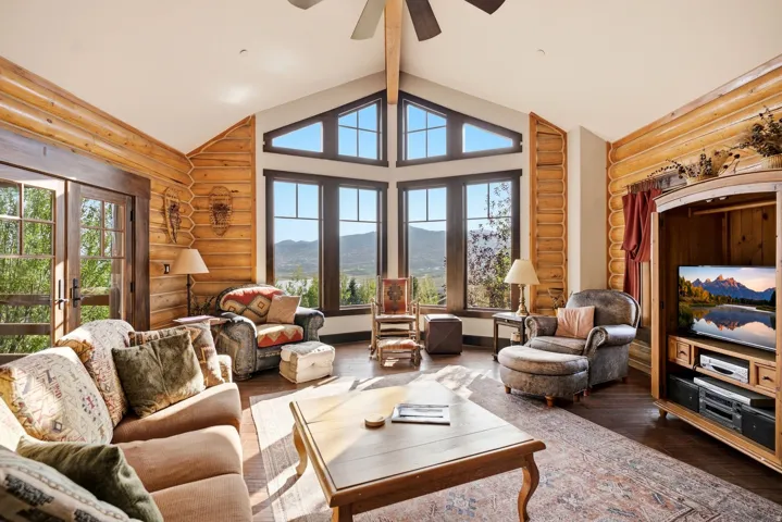 Vaulted ceiling with exposed beam, large picture windows, wood-finish flooring, log wall accents, and French doors