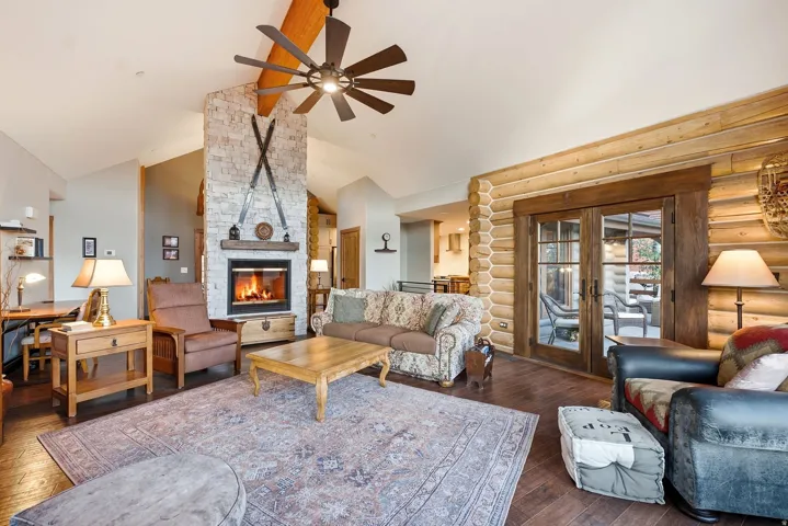 Stone fireplace with wood mantel, vaulted ceiling with exposed wood beam, wood-finish flooring, log wall accents, and double glass-paneled doors