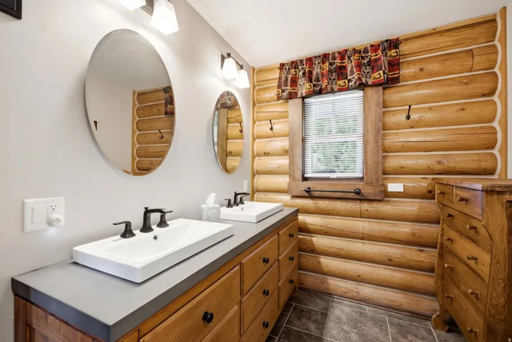 Dual vessel sinks with dark bronze fixtures, wood-finish vanity with storage, log-style wall paneling, and tile flooring