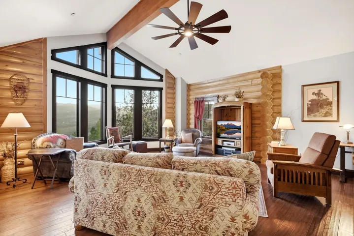 Vaulted ceiling with exposed wood beam, wood-finish flooring, custom log wall accents, multi-pane picture windows, and a ceiling fan