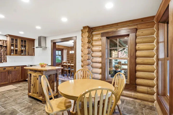 Wood-finish cabinetry with glass-front uppers, stainless steel range hood, stone-finish flooring, and log-style wall construction