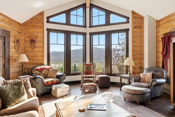 Vaulted ceiling with exposed wood beam, expansive windows with dark frames, wood-finish flooring, log wall accents, and an exterior door with dark wood trim