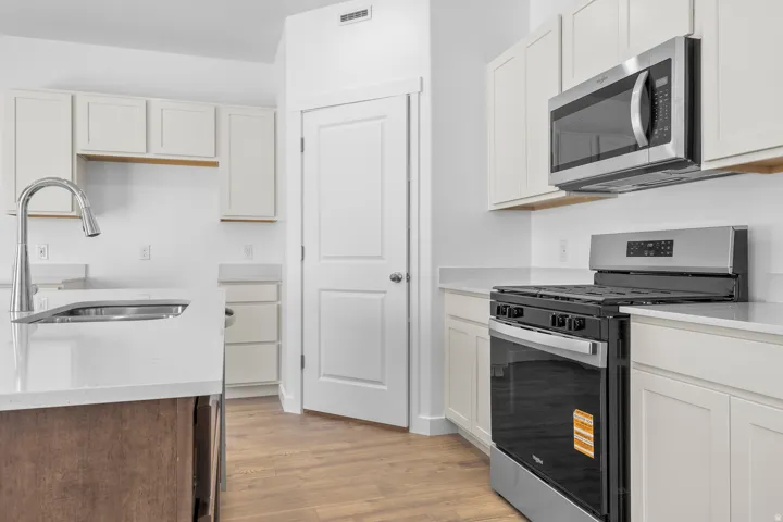 Kitchen featuring wood-finish flooring, white cabinetry, a kitchen island with an undermount sink, stainless steel appliances, and light-toned countertops