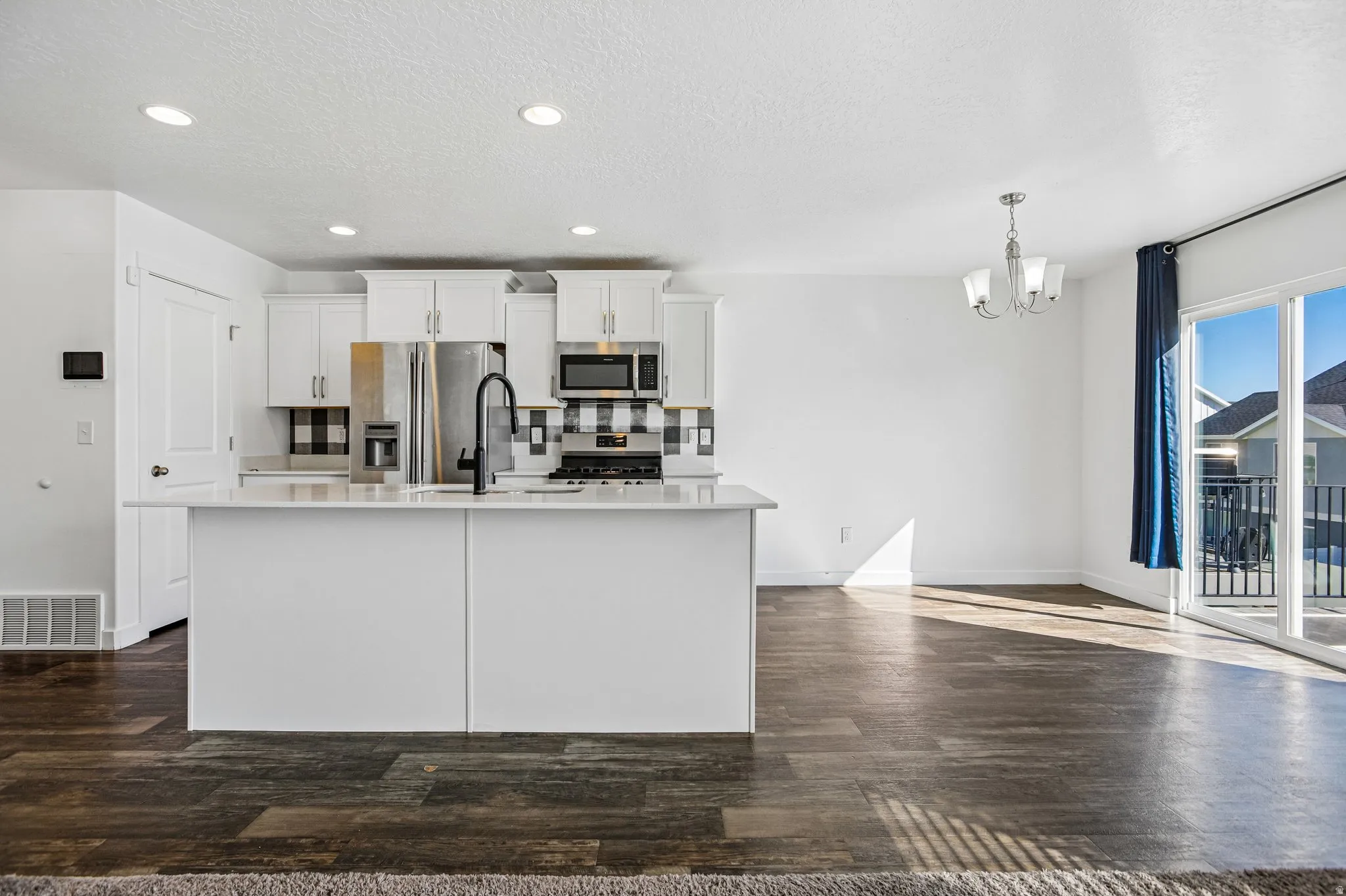 Open-concept living space with dark wood-finish flooring, white kitchen cabinetry, a central island with a sink, stainless steel appliances, and sliding glass doors