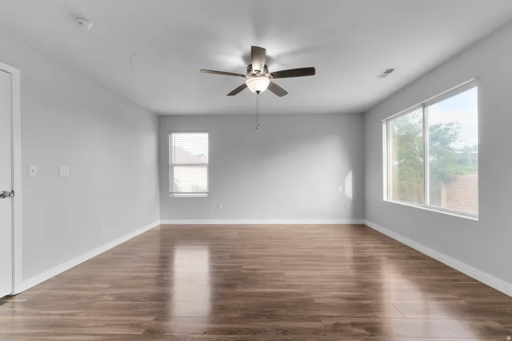Wood-finish flooring, ceiling fan with integrated lighting, two large windows, white walls, and a white door