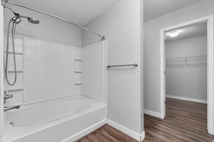 Bathtub with integrated shower, chrome-finish fixtures, white tile-patterned shower surround, wood-finish flooring, and a walk-in closet with wire shelving