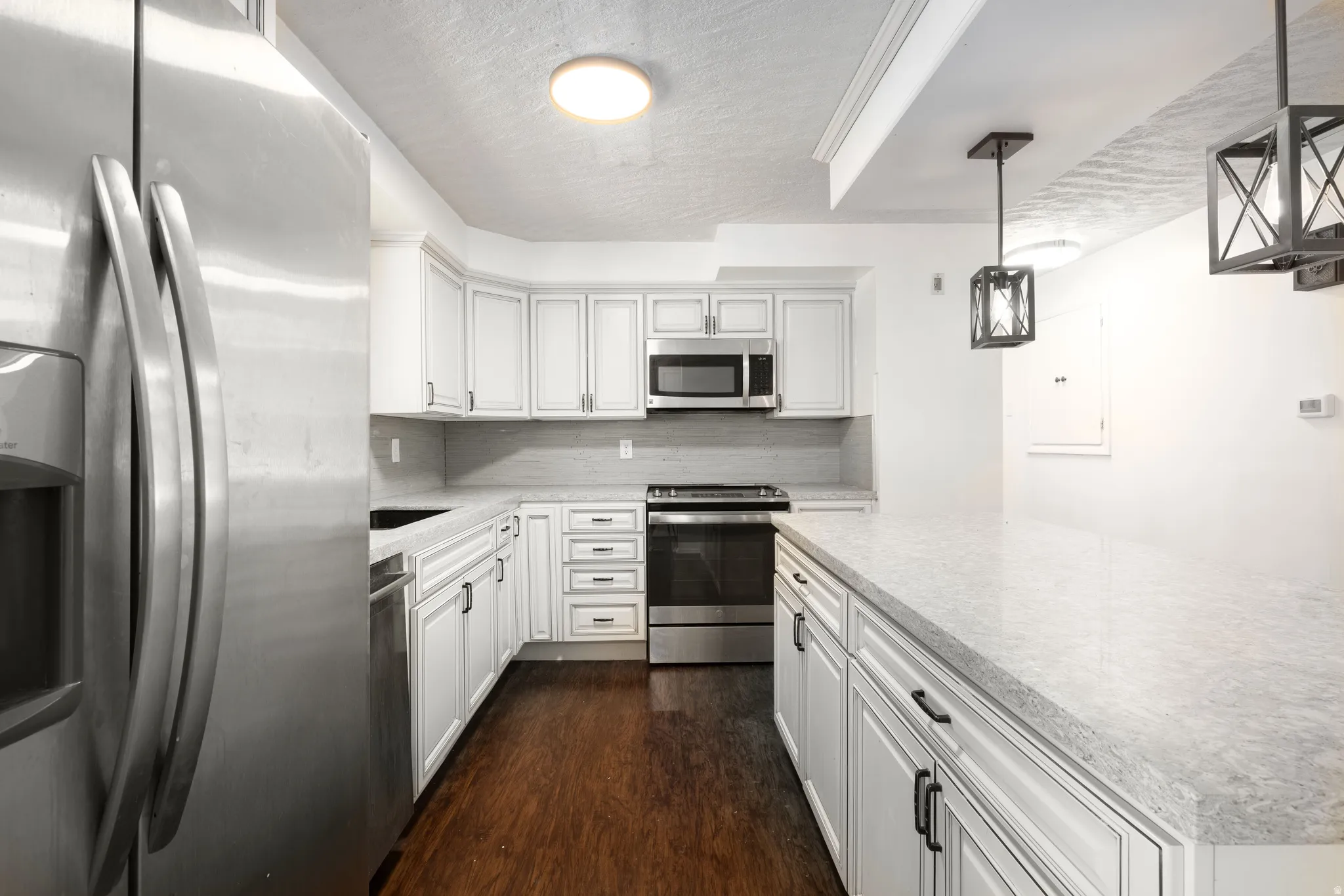 White cabinetry with dark hardware, stainless steel appliances, light-toned countertops, wood-finish flooring, and pendant lighting