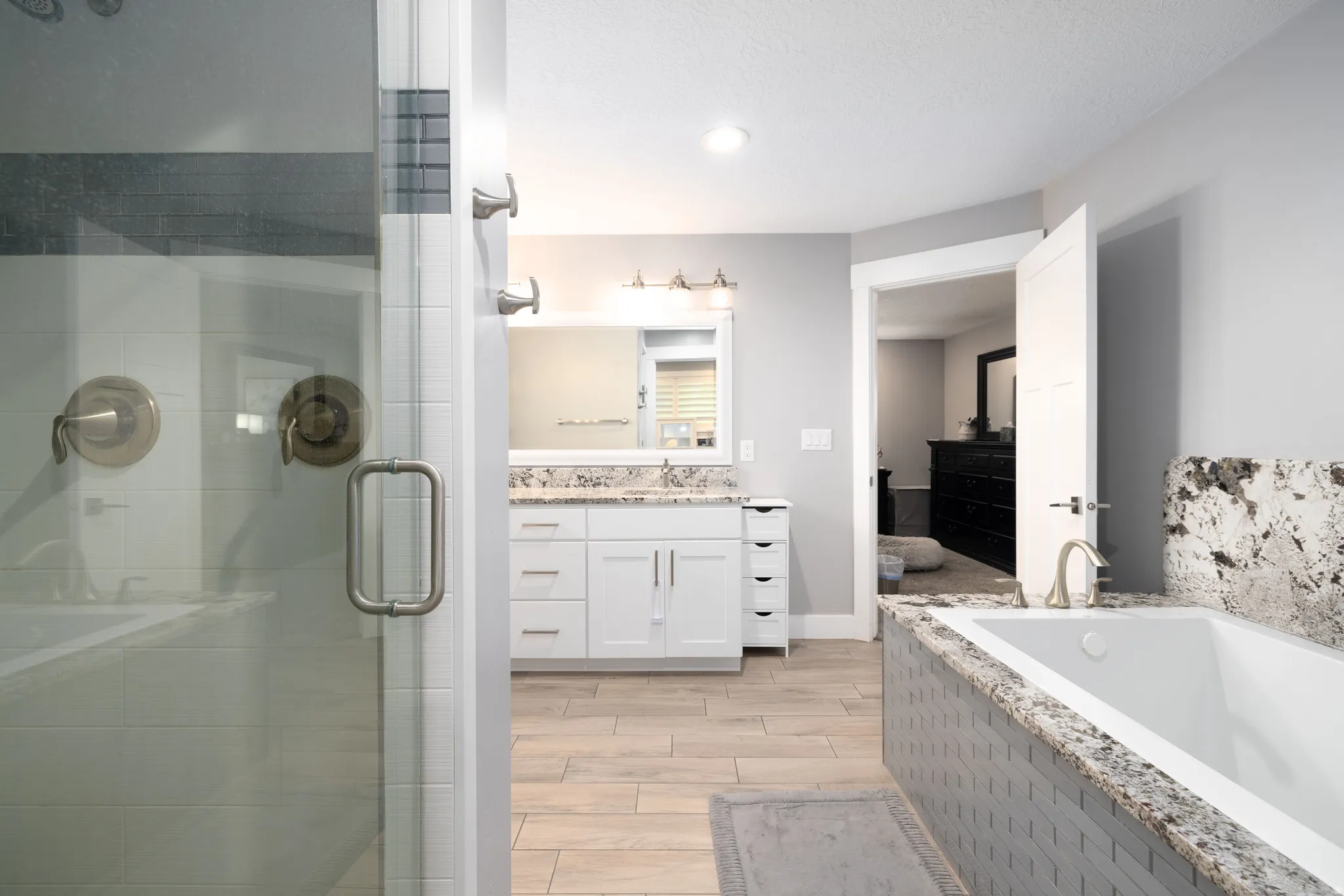Glass-enclosed shower with brushed metal fixtures, dual vanity with undermount sinks, built-in bathtub with tiled surround and stone ledge, and wood-finish flooring