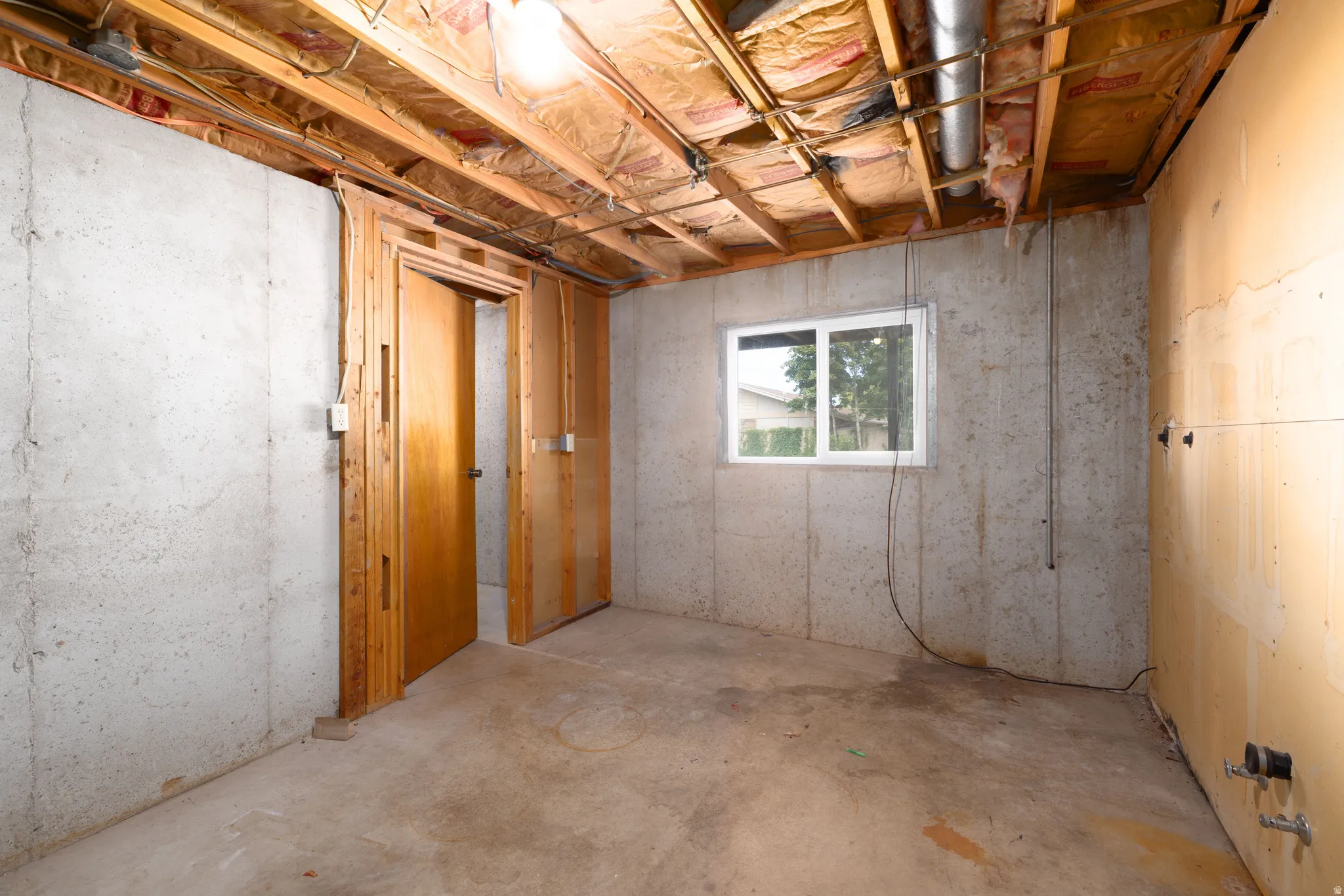 Exposed ceiling joists with insulation, concrete walls, single window with white frame, and two interior wooden doors