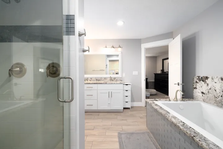 Glass-enclosed shower with brushed metal fixtures, dual vanity with undermount sinks, built-in bathtub with tiled surround and stone ledge, and wood-finish flooring